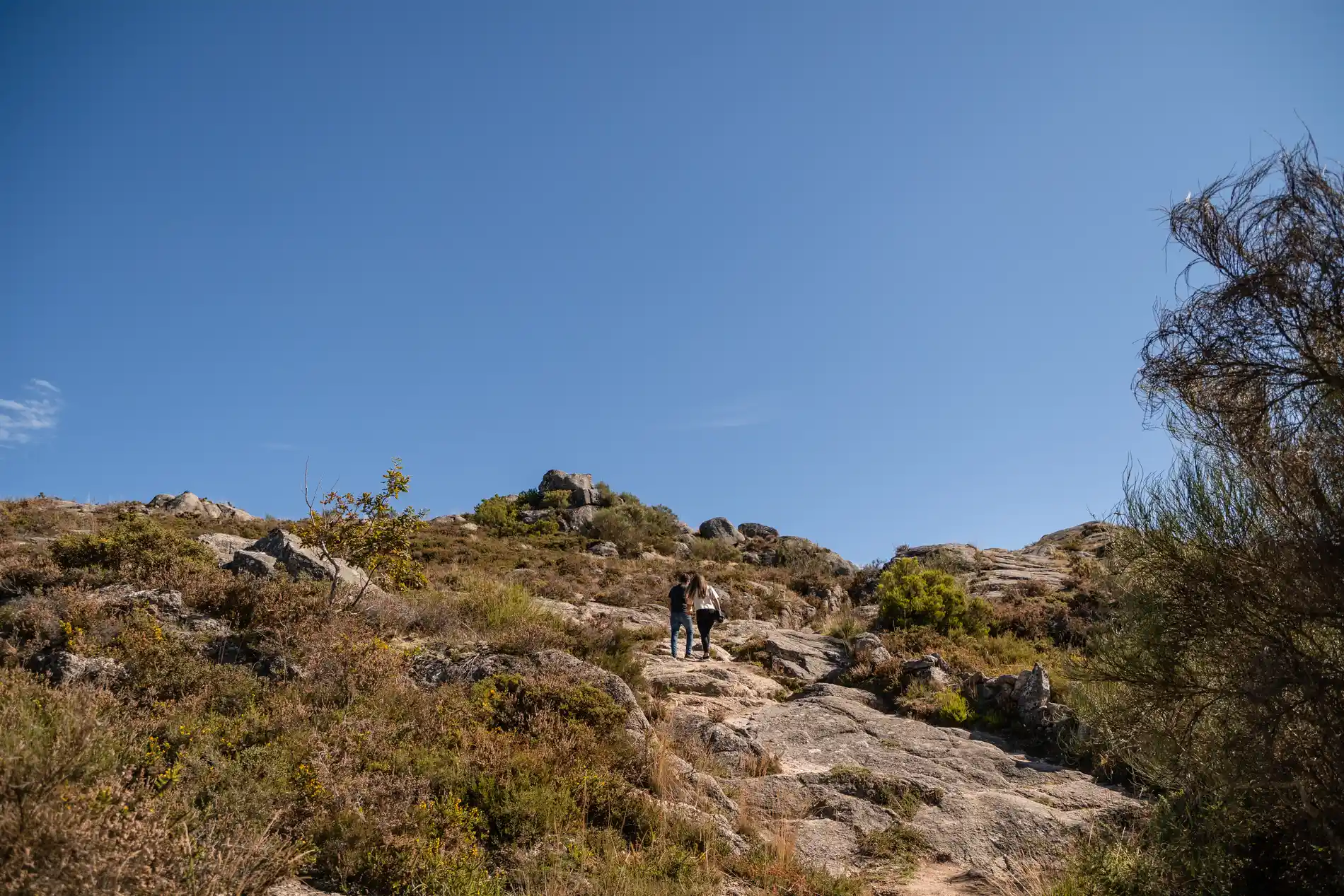 Trilha rochosa no campo; duas pessoas caminham sob céu azul