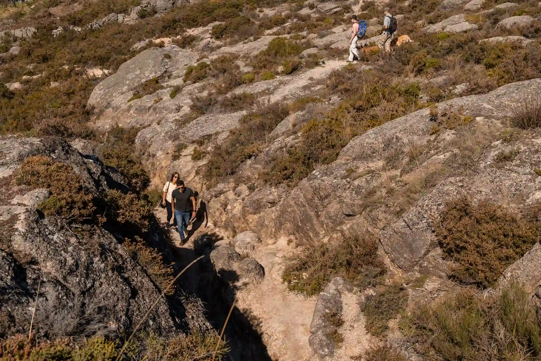 Caminhantes com mochilas em trilha rochosa, cruzando vale em montanha
