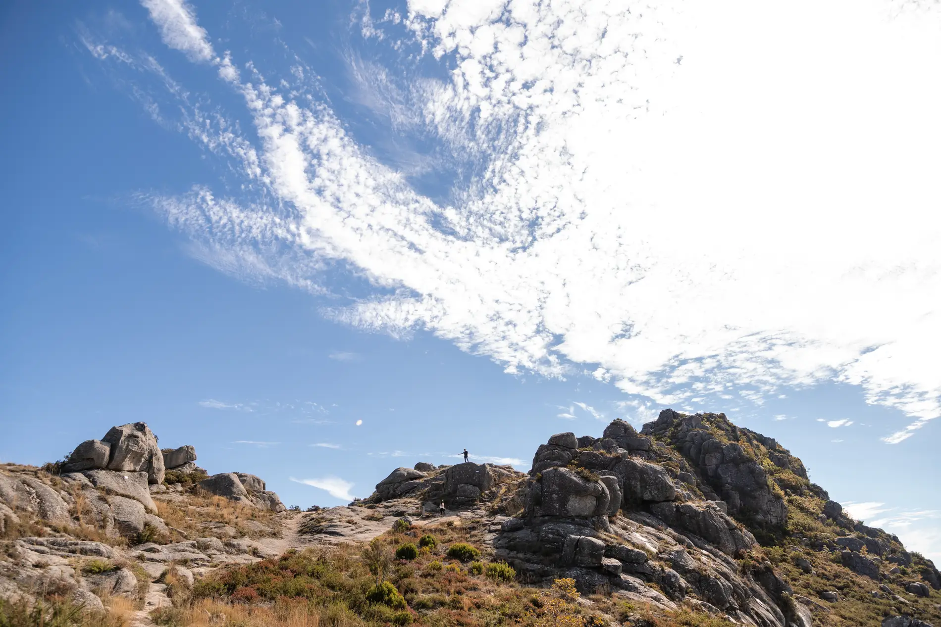 Céu azul e nuvens sobre trilha rochosa com pessoas no alto da montanha