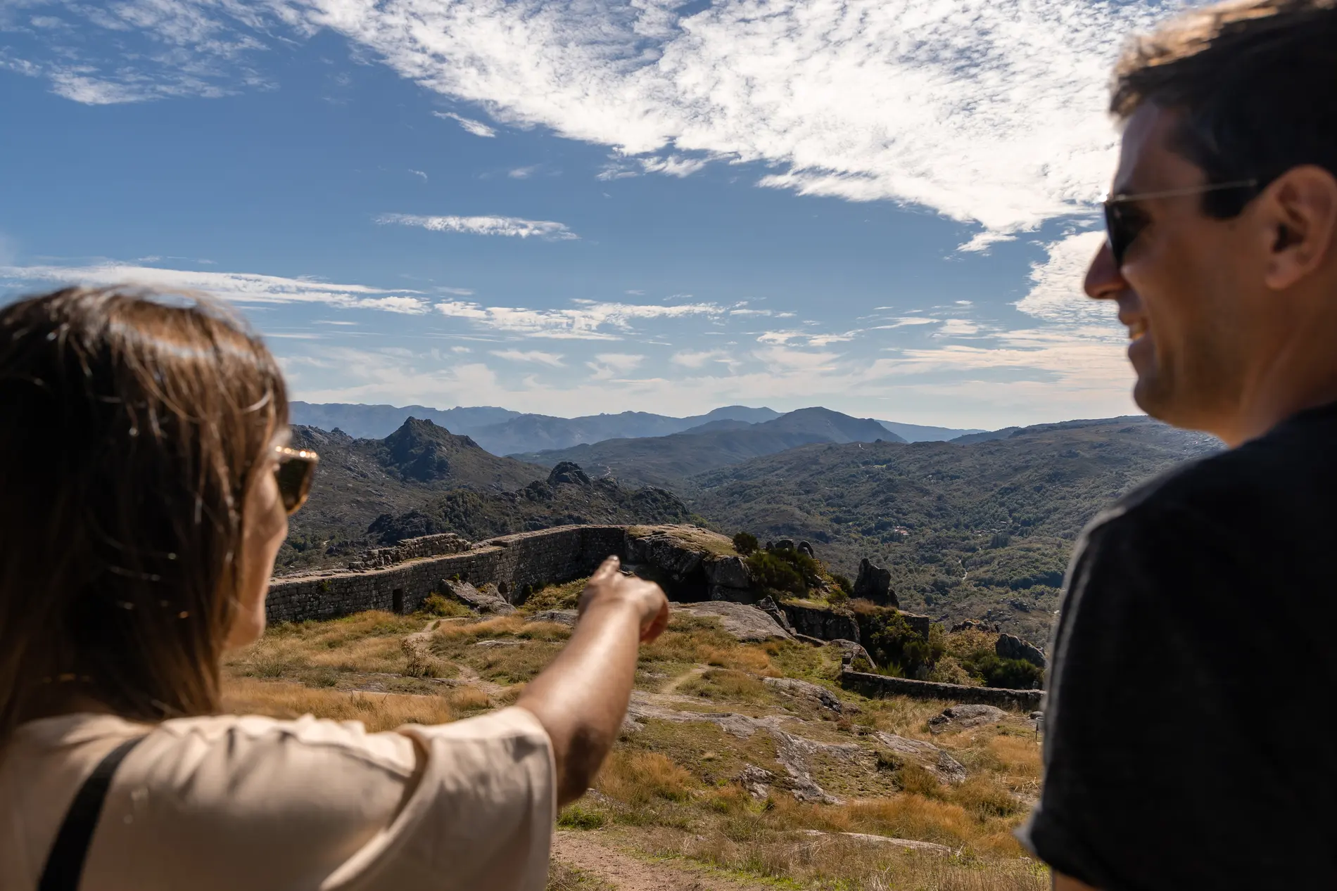 Turistas apontam para montanhas ao pôr do sol, com céu nublado e vales ao fundo