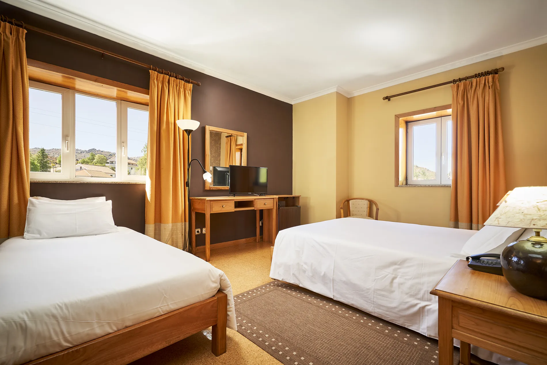 Quarto com duas camas, cortinas amarelas e mesa com TV e abajur