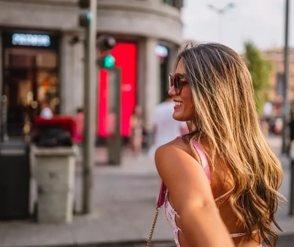 Mujer sonriente con gafas y cabello largo en una calle urbana soleada