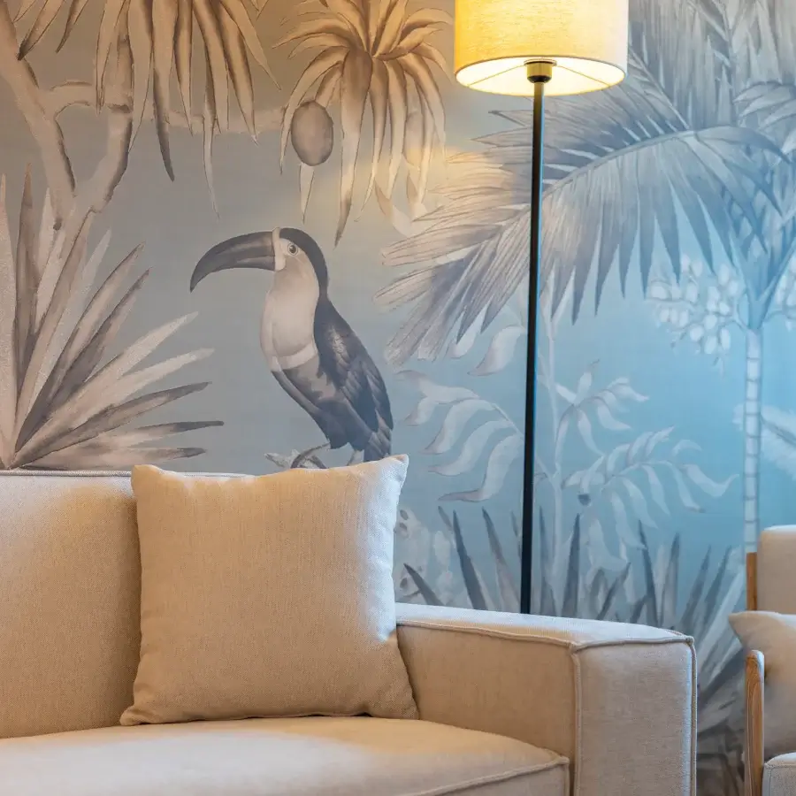 Tuc&aacute;n posado en una pared mural tropical junto a una l&aacute;mpara y sof&aacute; beige
