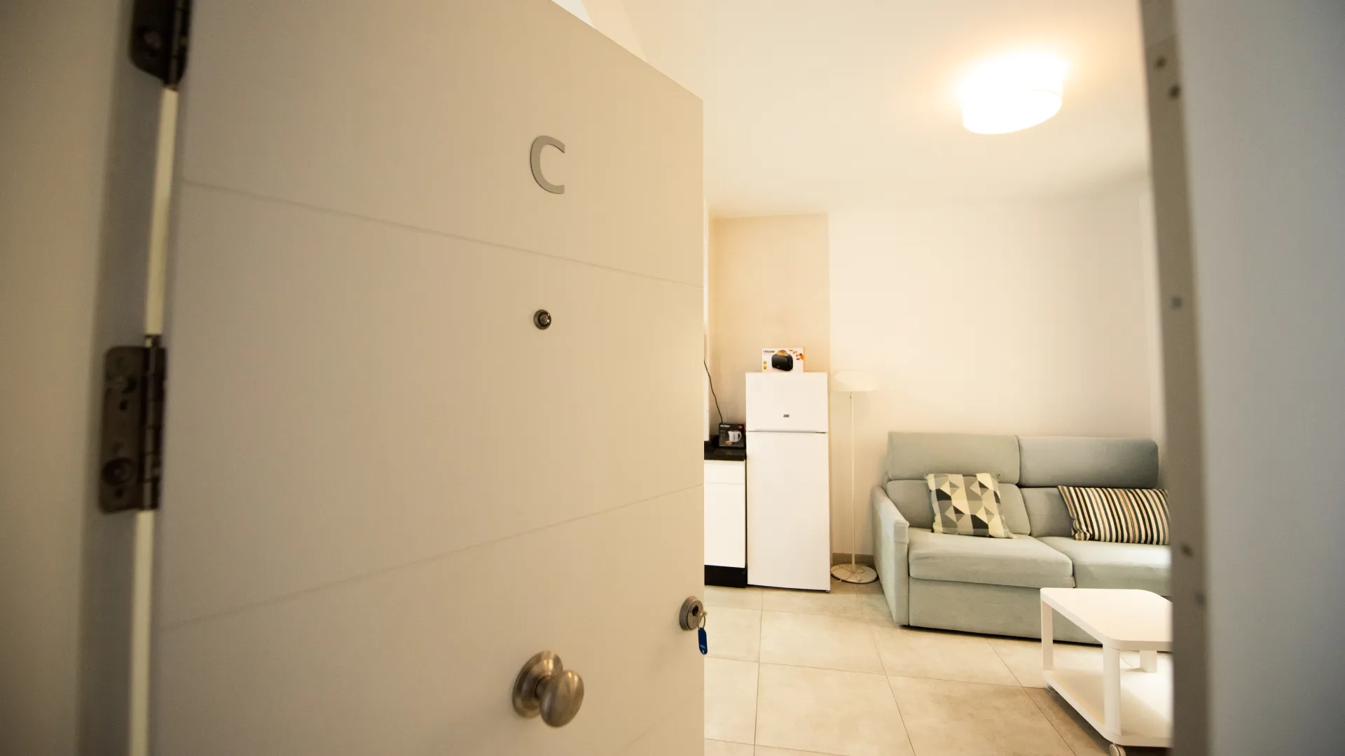Sala luminosa con sof&aacute; gris, cocina al fondo y puerta abierta.