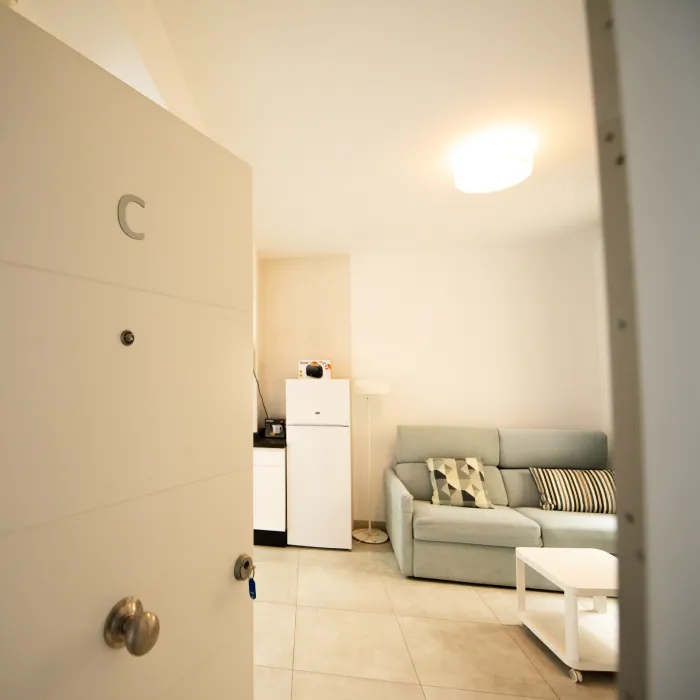 Sala con sof&aacute; claro, mesa blanca y nevera al fondo, vista desde la puerta
