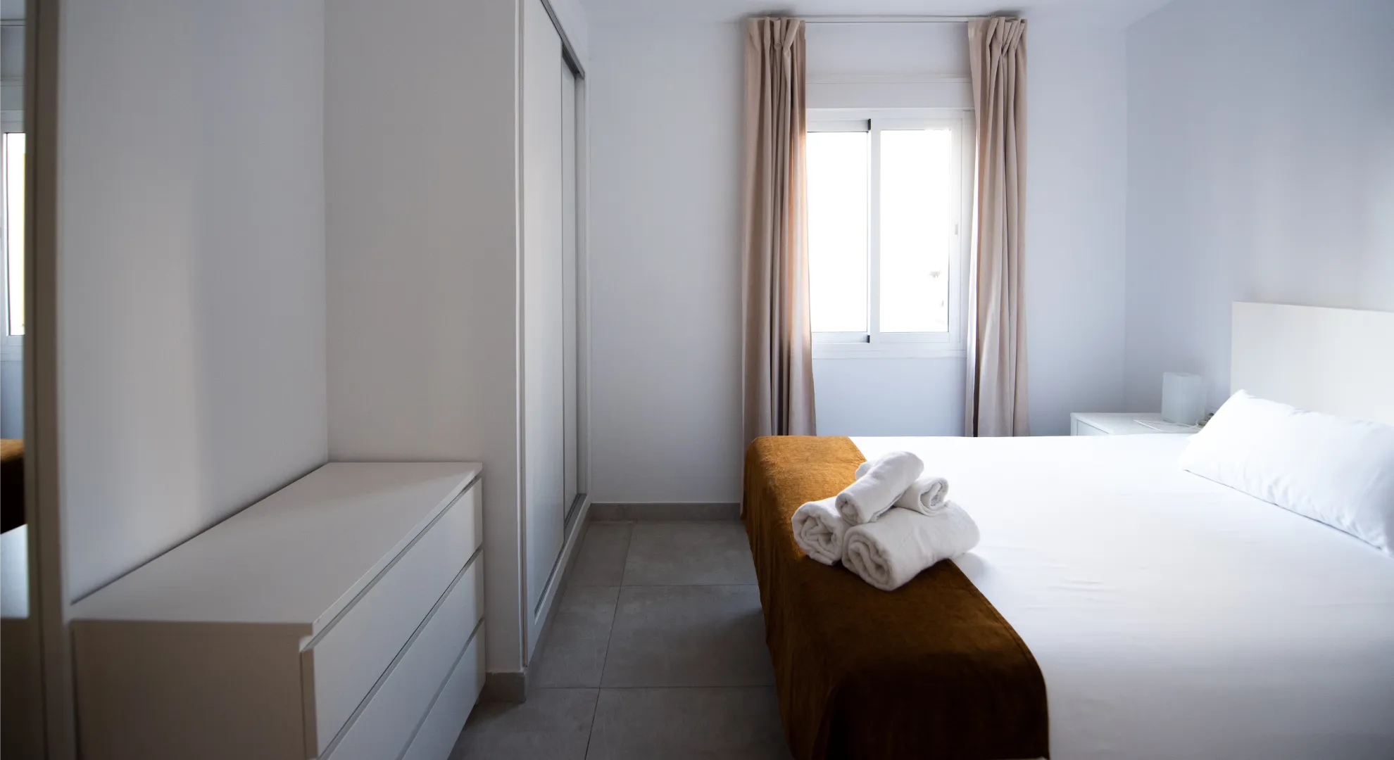 Dormitorio moderno con cama blanca, cortinas beige y ventana al fondo