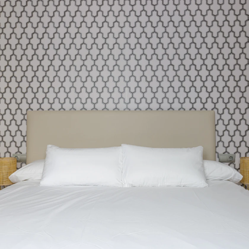 Cama con ropa blanca y dos cojines sobre fondo de pared con patr&oacute;n geom&eacute;trico