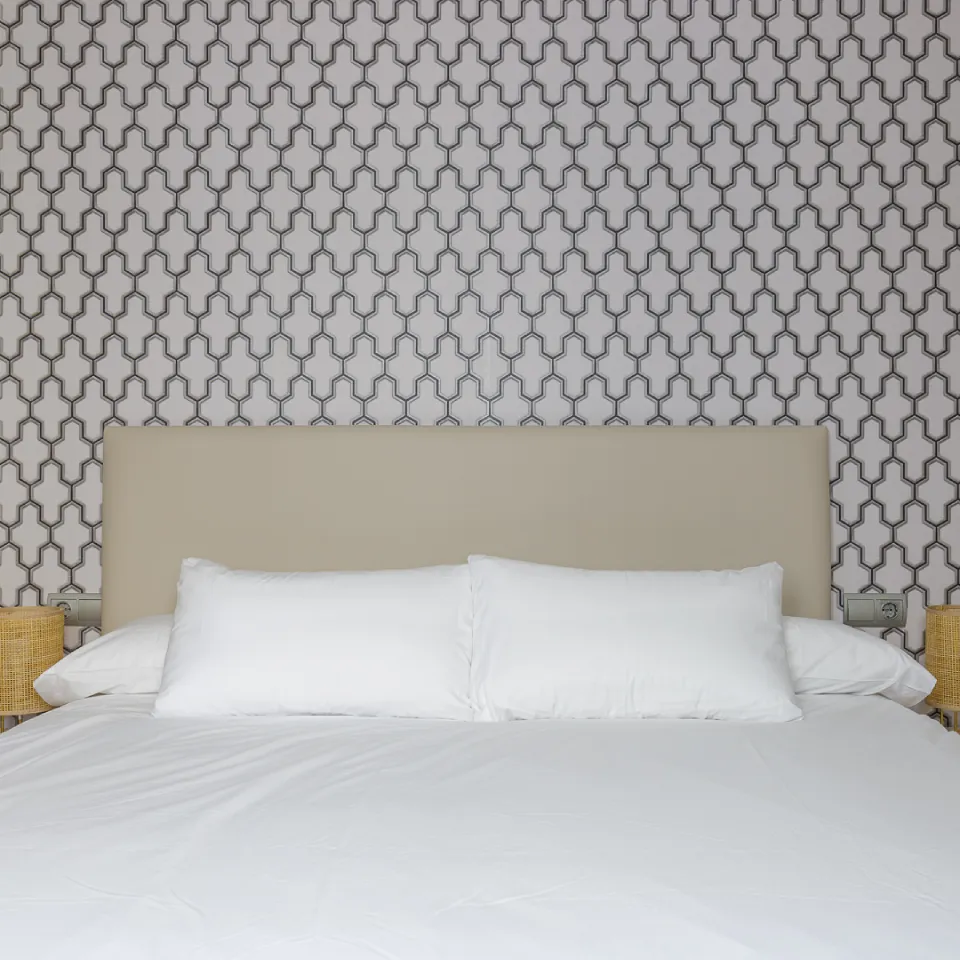Cama con cabecero beige y s&aacute;banas blancas en pared con patr&oacute;n geom&eacute;trico.