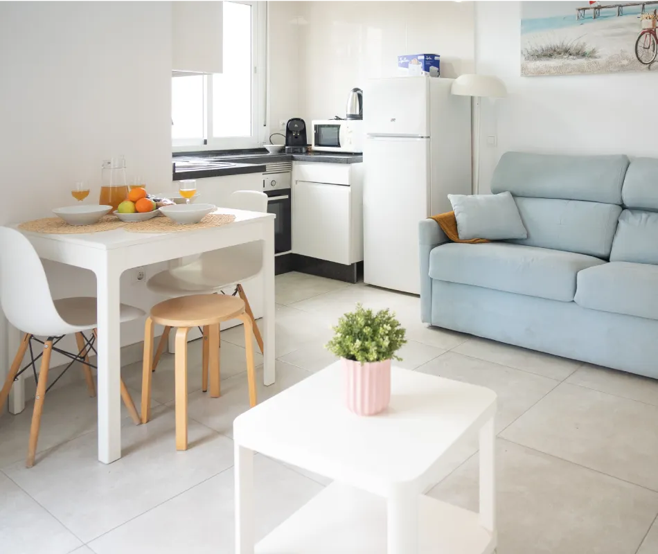Cocina y sala con sof&aacute; azul, mesa blanca y mesa de comedor con frutas