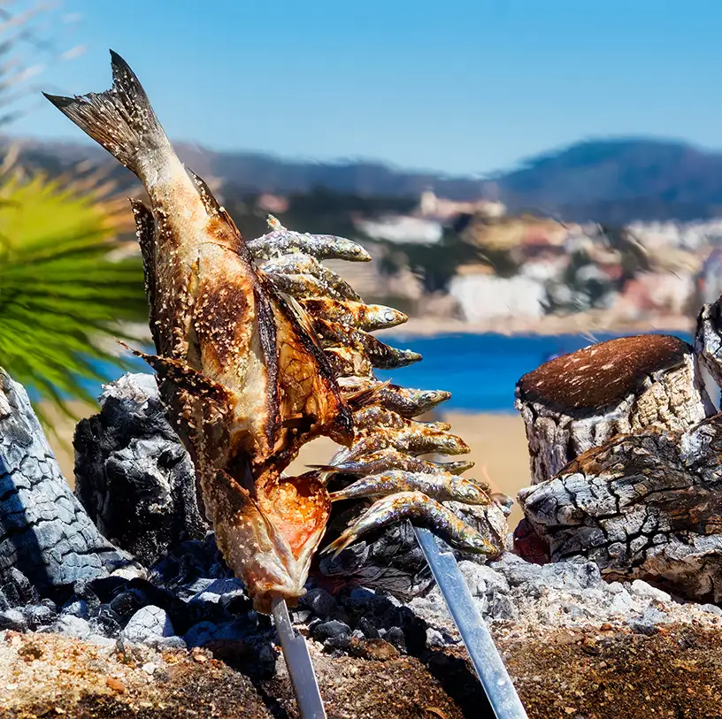Peces y sardinas as&aacute;ndose sobre brasas y madera, con ciudad al fondo.
