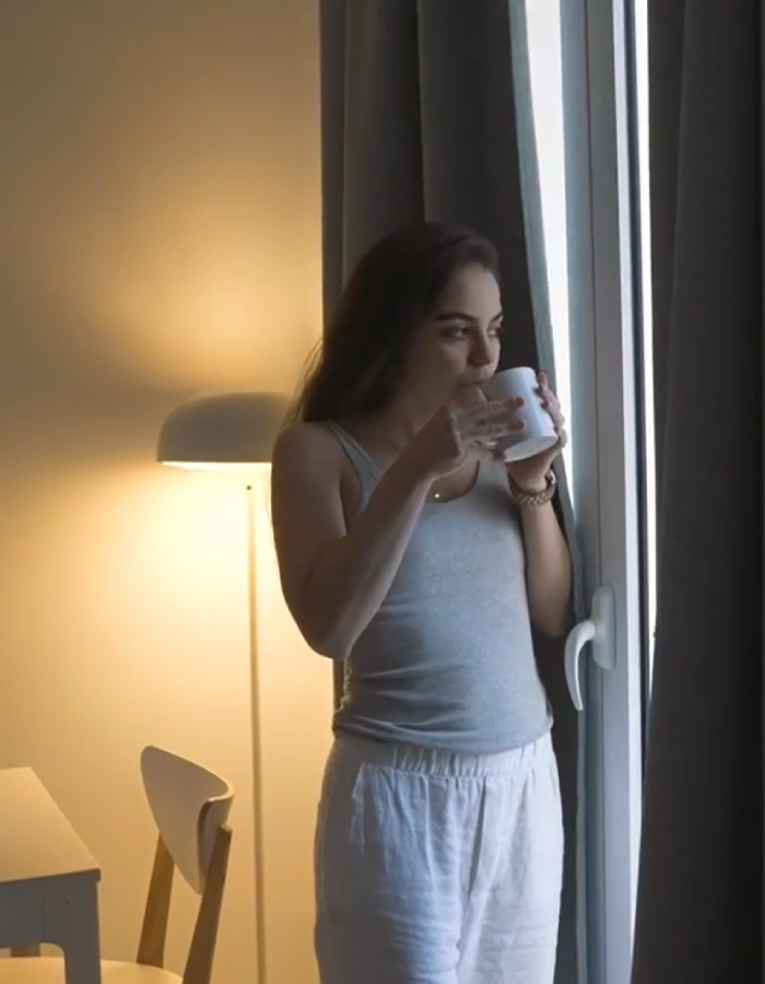 Mujer de pie junto a una ventana, bebiendo de una taza blanca.