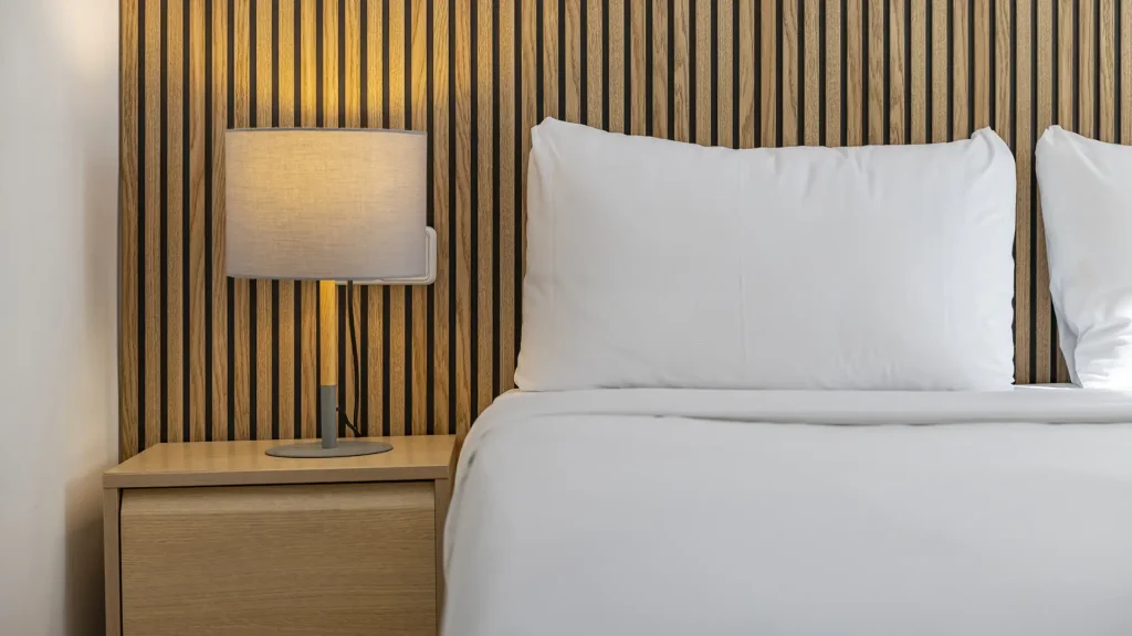 Cama con almohadas blancas y l&aacute;mpara sobre mesita de madera, pared de listones