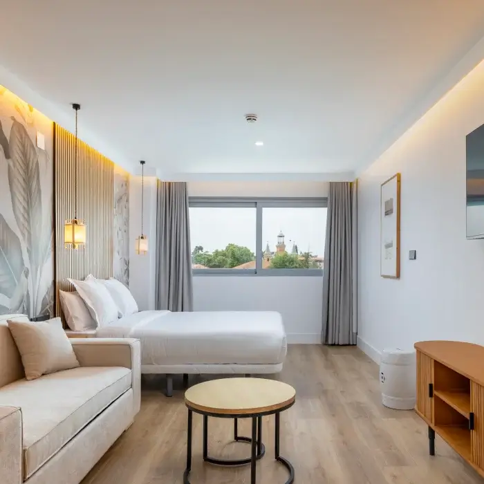 Dormitorio moderno con cama, sof&aacute;, cortinas grises y vista exterior.