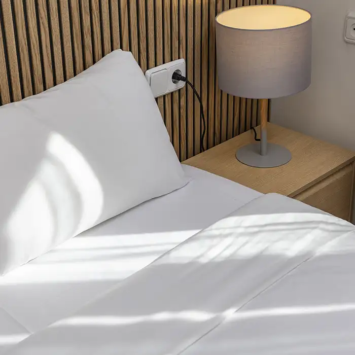 Cama con ropa blanca y l&aacute;mpara sobre mesa de madera, pared con listones.