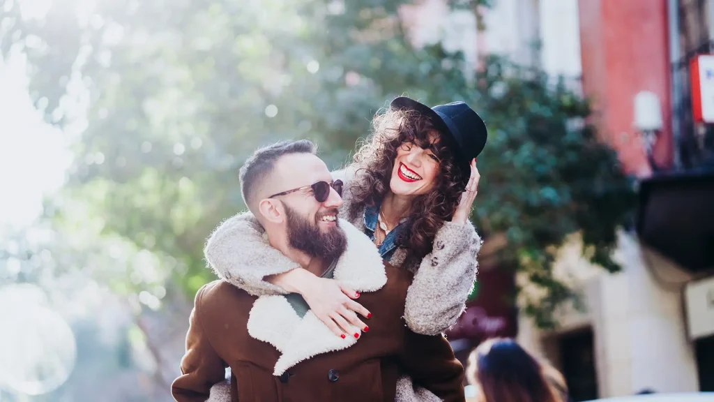 Pareja sonriente abrazada, ella con sombrero y pelo rizado.
