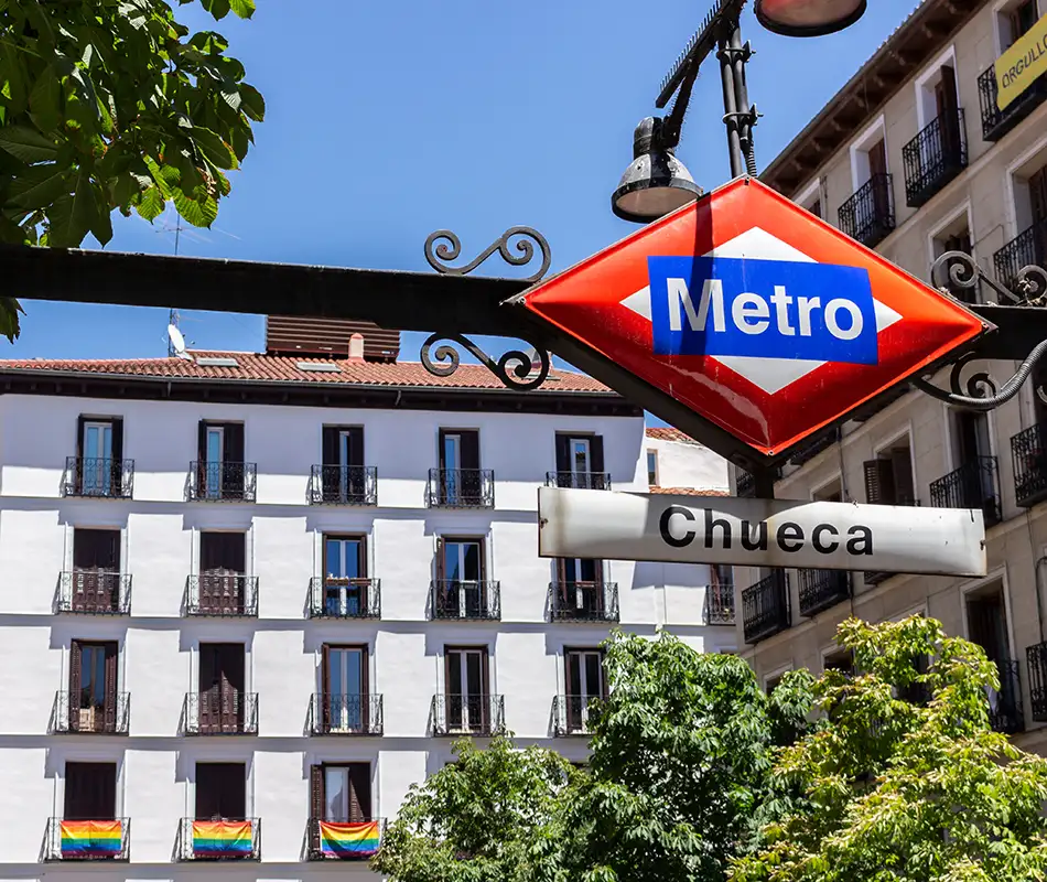 Se&ntilde;al de Metro &ldquo;Chueca&rdquo; en una calle de Madrid, con edificios y banderas.