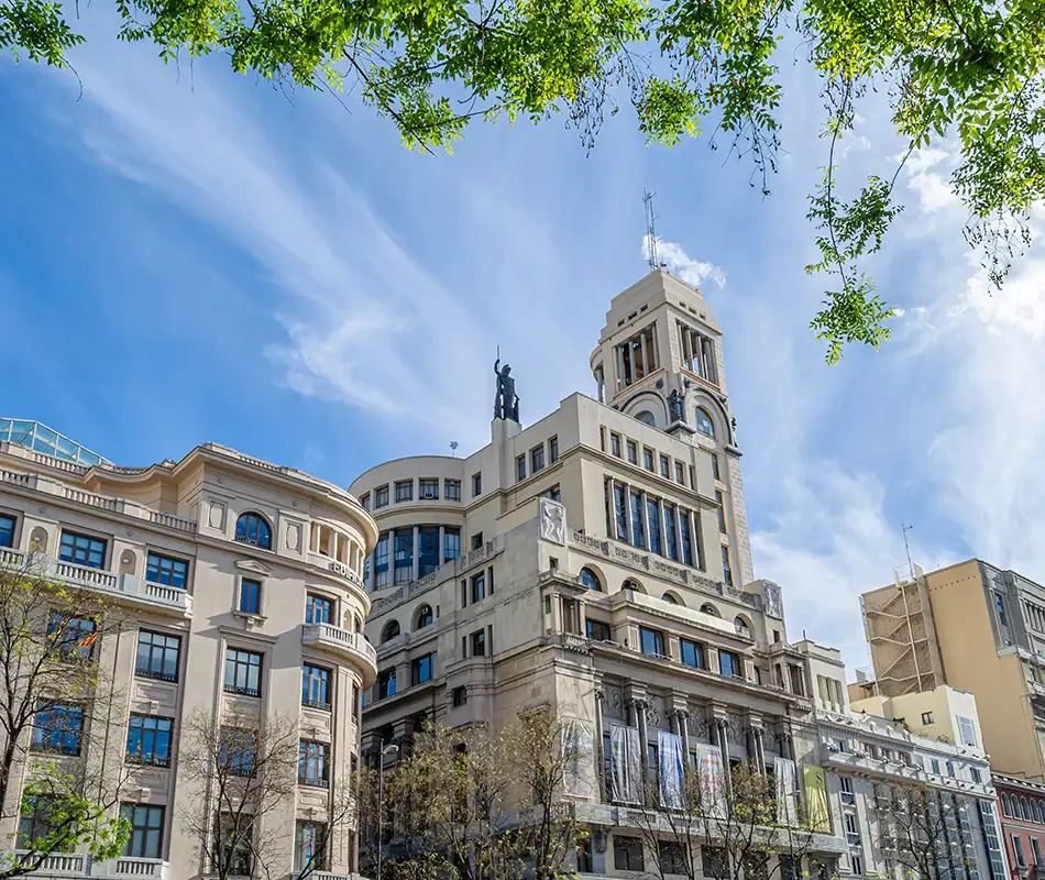 Edificio hist&oacute;rico con torre y estatua, cielo azul entre &aacute;rboles y nubes