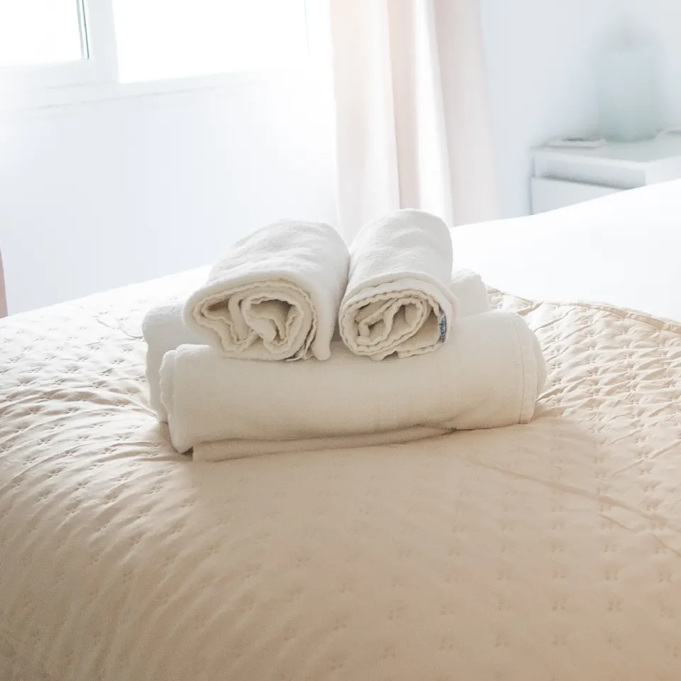 Rollo de mantas blancas dobladas sobre una cama de dormitorio, en el fondo cortinas.