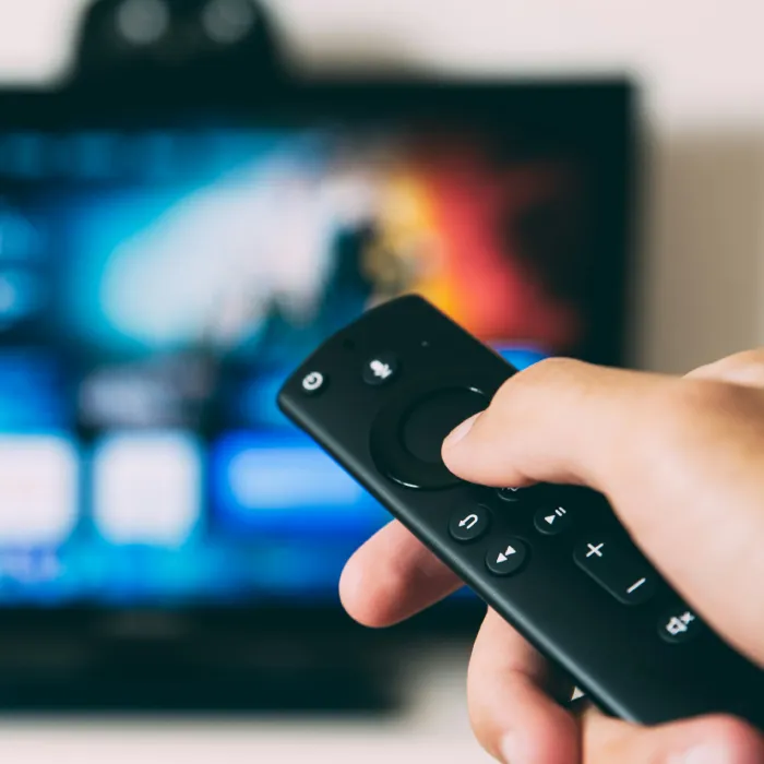 Mano sosteniendo un control remoto negro frente a una TV con fondo desenfocado
