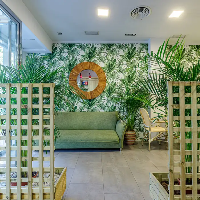 Sala con sof&aacute; verde y pared con hojas tropicales, espejo redondo y luz.