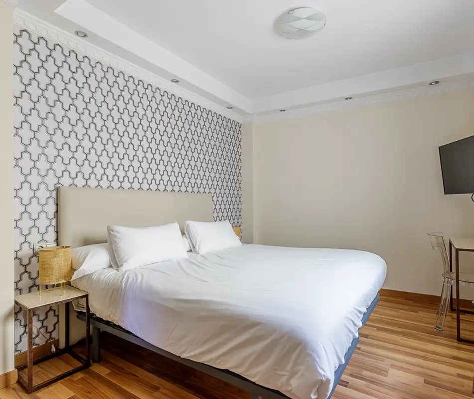 Dormitorio moderno con cama blanca, pared geom&eacute;trica y luz de techo.