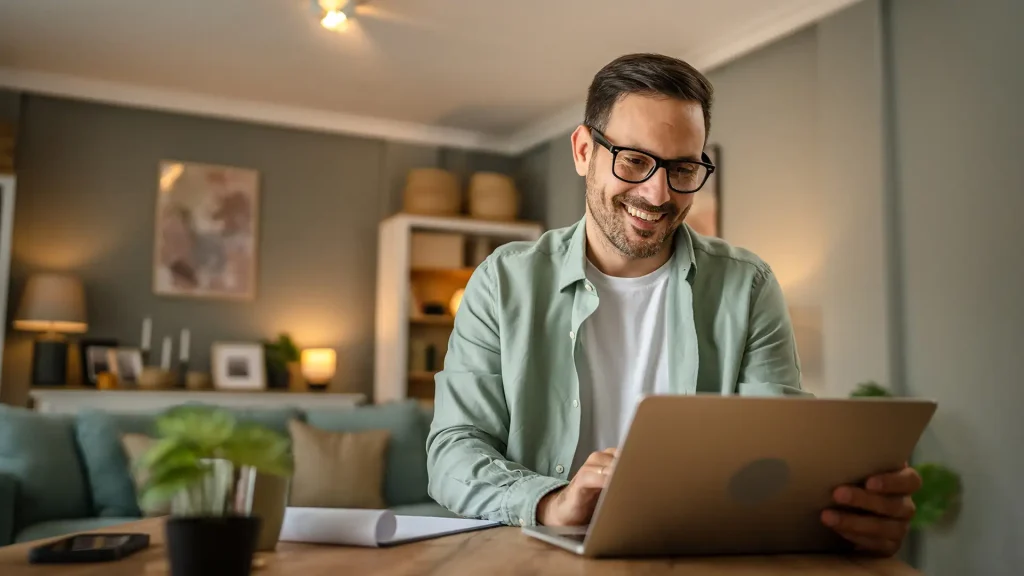 Hombre con gafas sonr&iacute;e frente a una computadora port&aacute;til en casa