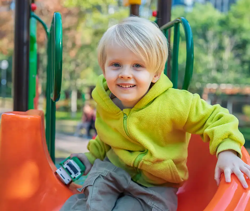 Ni&ntilde;o sonriente en un parque infantil con ropa verde y tobog&aacute;n naranja.