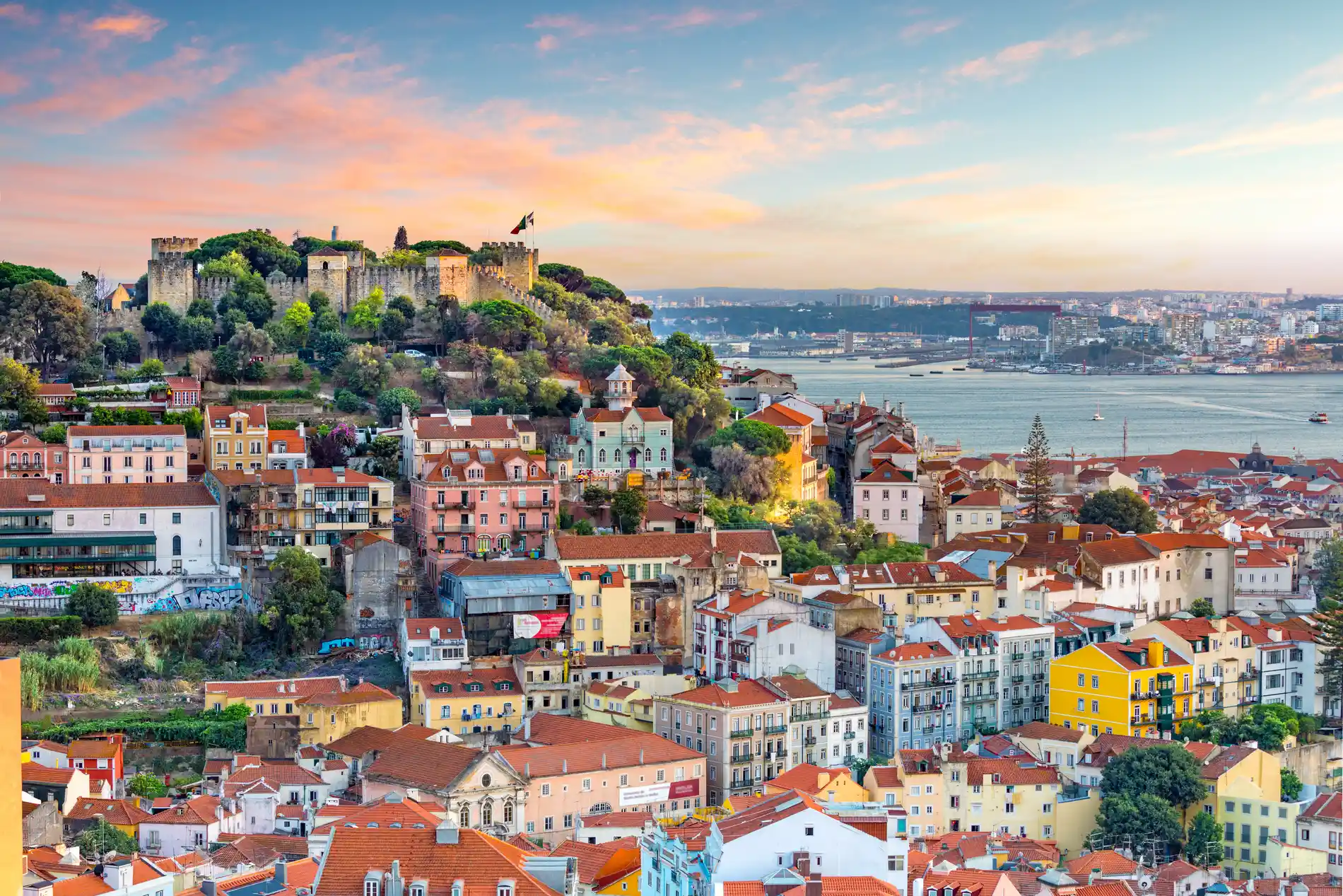 Vista panor&acirc;mica de Lisboa com muralhas antigas, casas coloridas e rio ao p&ocirc;r do sol