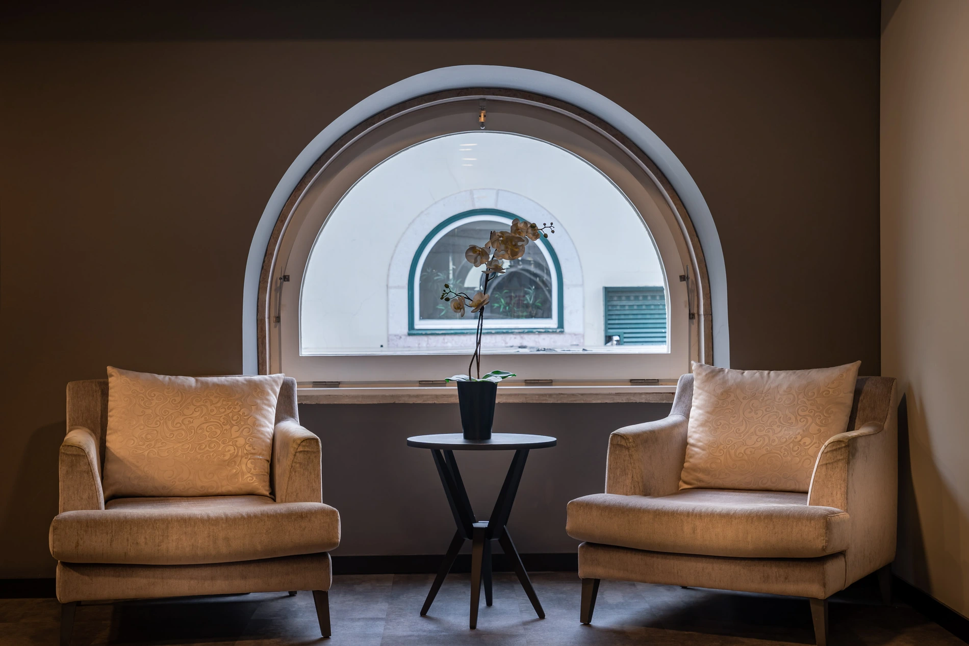 Sala com duas poltronas e mesa, janela circular com plantas ao centro