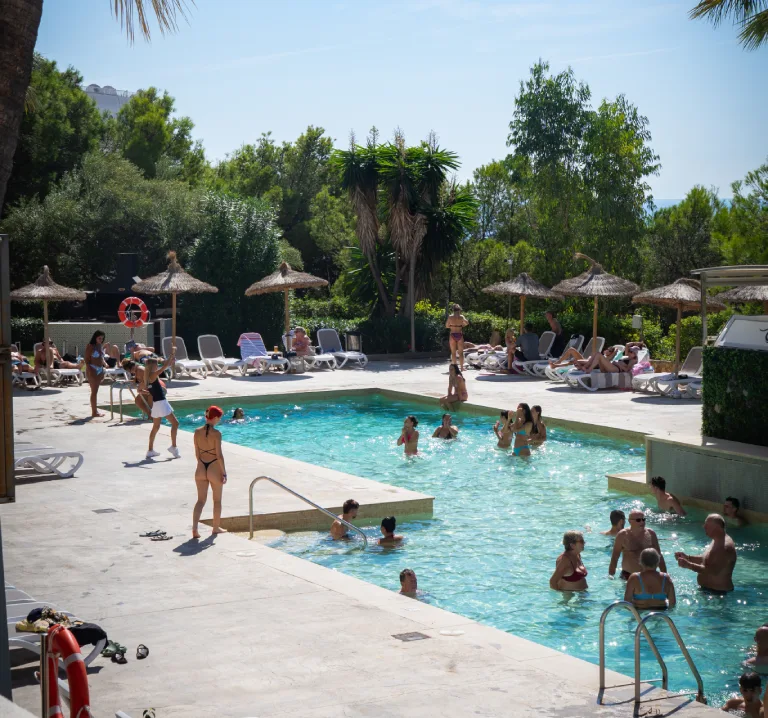 Piscina con ba&ntilde;istas y tumbonas bajo palmeras, en un resort al sol