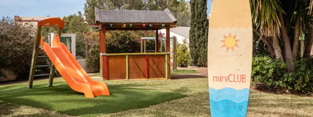 Parque infantil con tobog&aacute;n naranja y mural miniCLUB, jard&iacute;n soleado