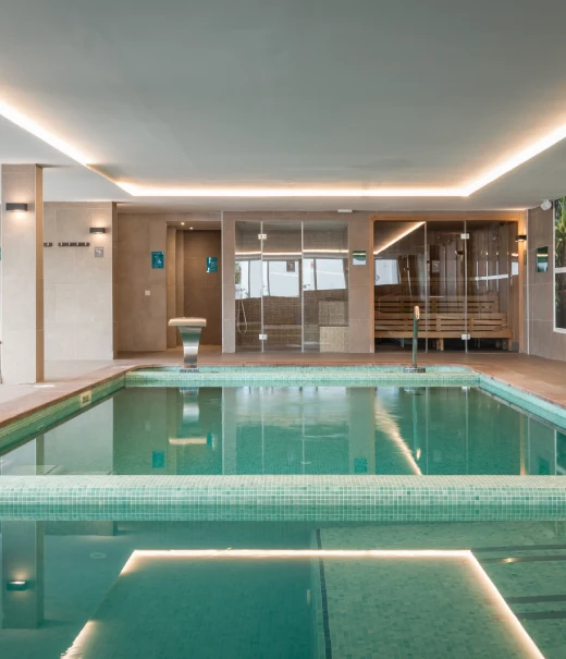 Piscina interior con iluminaci&oacute;n LED y zona de descanso al fondo, spa.