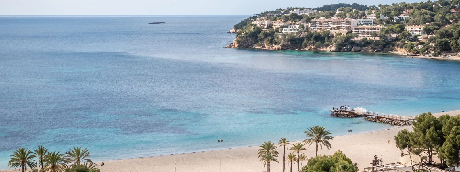 Playa de aguas turquesas con paseo mar&iacute;timo y acantilados al fondo.