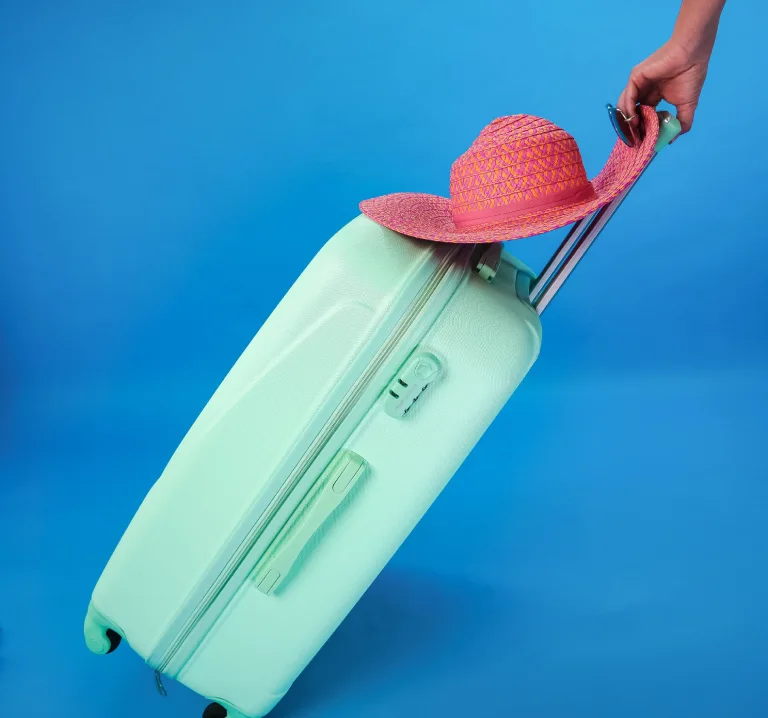 Mano con sombrero de paja sobre maleta de viaje en fondo azul