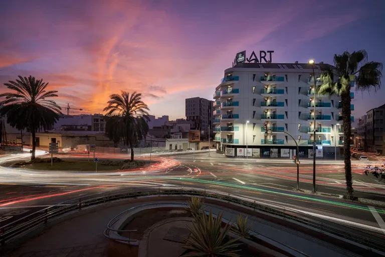 Hotel moderno con palmeras y luces de tr&aacute;fico al atardecer