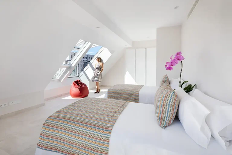 Mujer en habitaci&oacute;n luminosa con cama blanca y flores fucsia decorativas