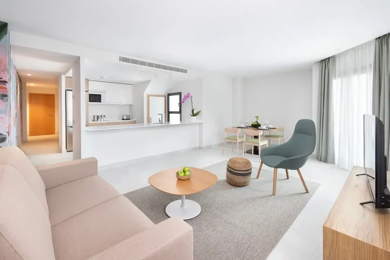 Sala de estar moderna blanca, con sof&aacute; beige, mesa y mesa auxiliar junto a ventanales