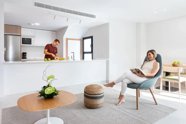 Mujer leyendo en una sala luminosa, cocina con hombre preparando alimentos.