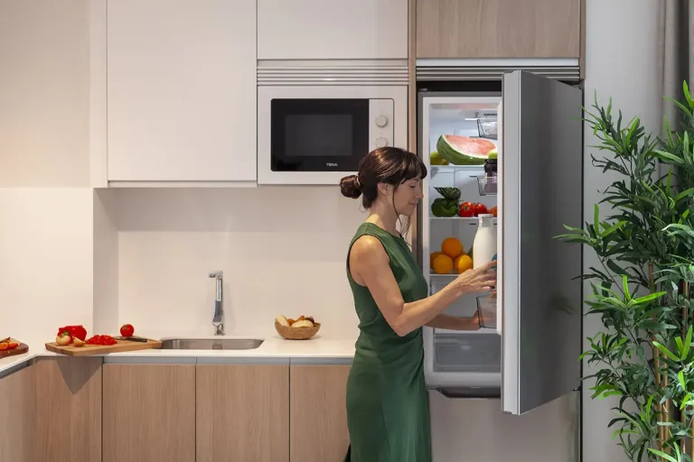 Mujer prepara comida frente a la puerta del frigor&iacute;fico abierto en cocina moderna.