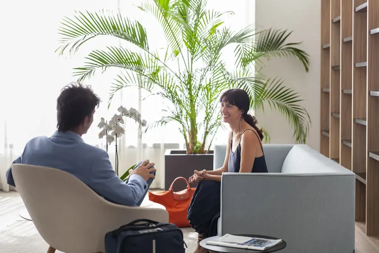 Dos personas conversan sentadas en una oficina moderna junto a una planta.
