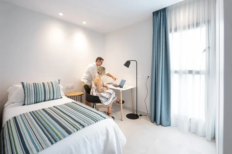 Habitaci&oacute;n luminosa con cama, mesa de trabajo y padre ayudando a una ni&ntilde;a