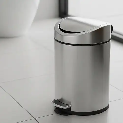 Papelera met&aacute;lica gris con pedal y tapa abatible en un suelo blanco.