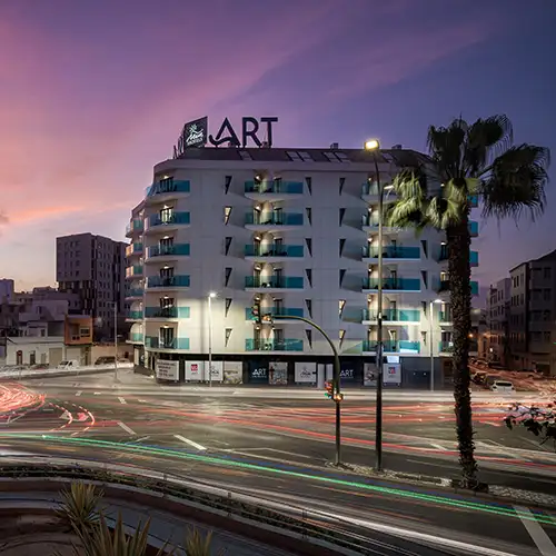 Edificio &ldquo;ART&rdquo; iluminado al atardecer, con palmeras y tr&aacute;fico de noche