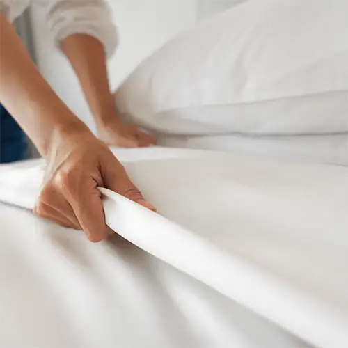 Manos doblando una s&aacute;bana blanca sobre una cama