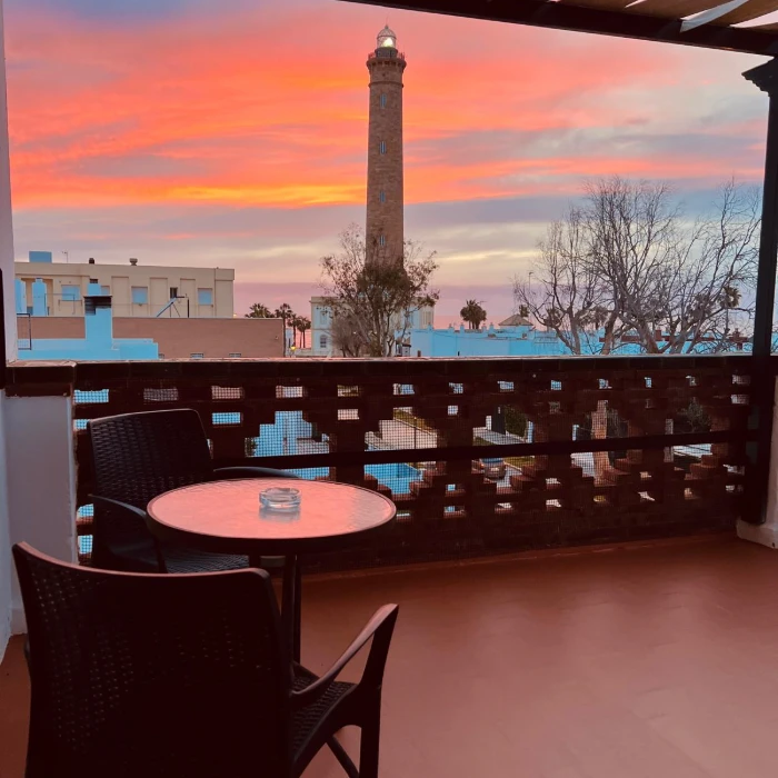 Terraza con mesa y sillas frente a un faro en un atardecer rosado.