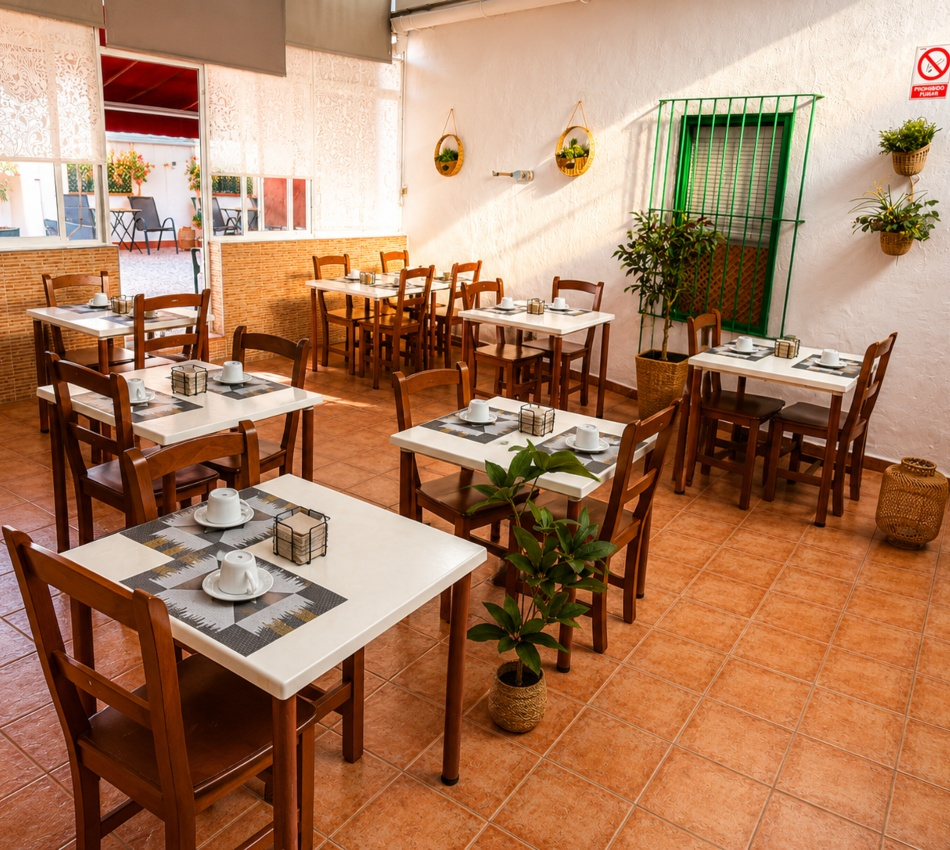 Comedor con mesas y sillas de madera, plantas y ventana en pared blanca.