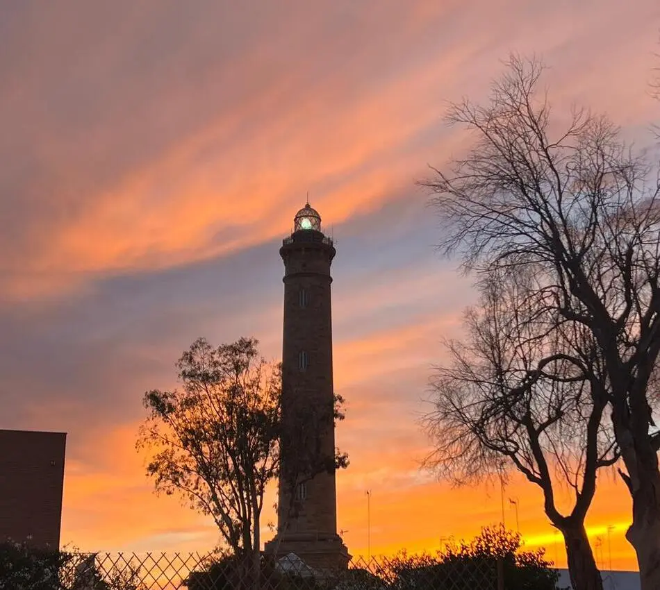 Faro junto a &aacute;rboles en un atardecer naranja con cielo nublado