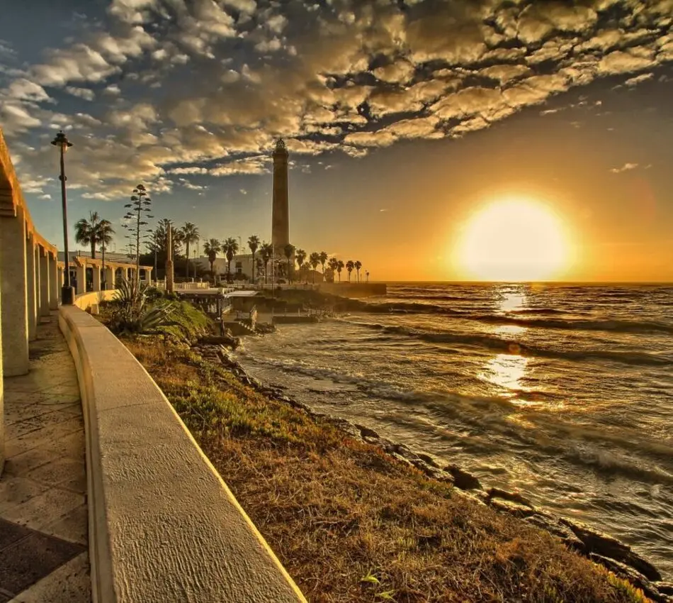 Puesta de sol en la costa con palmeras, oleaje y un faro al fondo.
