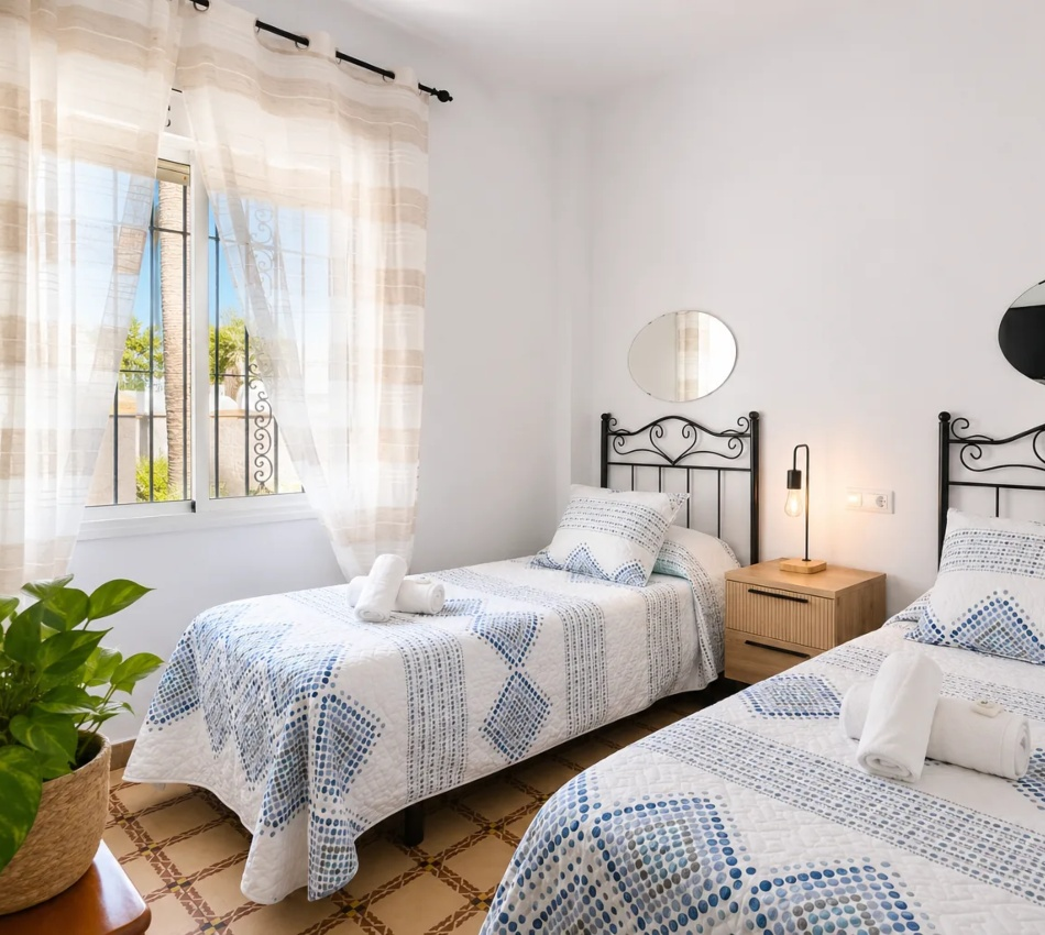 Dormitorio con dos camas, cortinas blancas y cojines azules, con espejo ovalado
