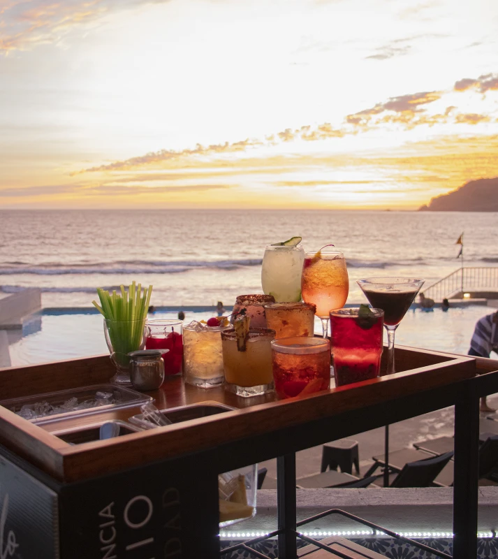 Bandeja con c&oacute;cteles y cena frente al mar al atardecer.